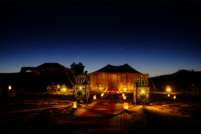 A beautifully lit desert camp at night, surrounded by sand dunes under a starry sky