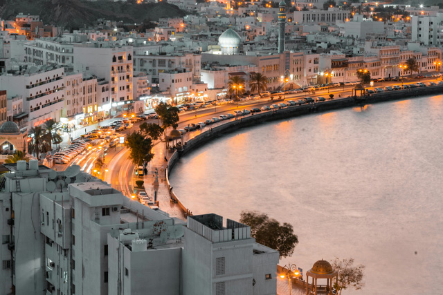A stunning evening view of Mutrah Corniche in Muscat, Oman, with a curved waterfront, illuminated streets, and traditional white buildings.